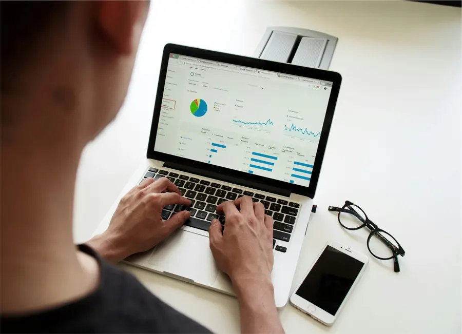 Person typing on a laptop showing analytics charts, with glasses and a phone beside it