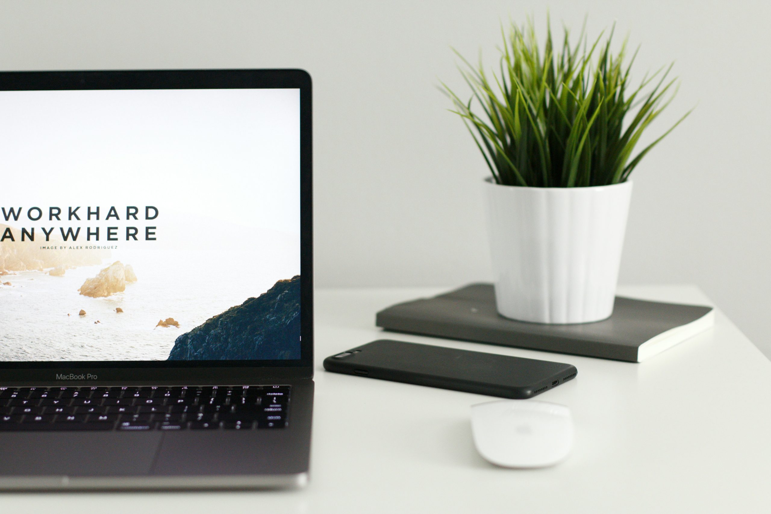 MacBook Pro displaying "Work Hard Anywhere" on screen, alongside a black smartphone, a notebook, and a white planter with green grass, symbolising remote work and website performance optimisation.